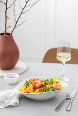 Elegante Pastaschale mit frischem Salat und Garnelen, seitliche Perspektive, auf einem gedeckten Tisch mit Weinglas und Vase im Hintergrund.