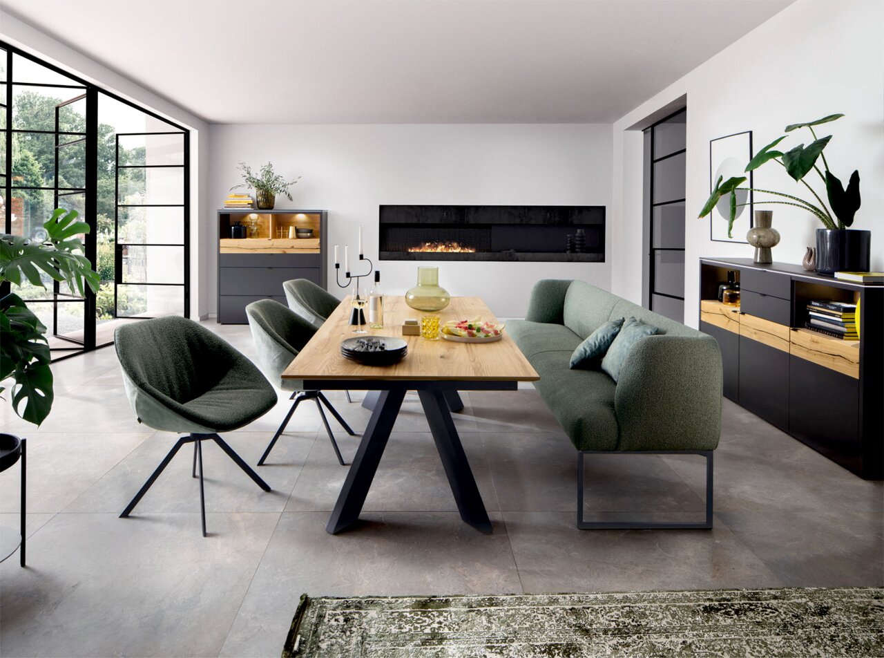 Moderne eetkamer met houten eettafel op zwart metalen onderstel, groene gestoffeerde stoelen en bank, en zwarte dressoirs met houtaccenten; perspectief: frontale, licht schuin van links genomen kameropname.