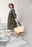 Frau in einem olivgrünen Trenchcoat, die eine mittelgroße schwarze Shopper-Tasche aus Raffia-Material trägt, während sie eine Treppe hinuntergeht. Seitenansicht.
