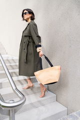 Frau in einem olivgrünen Trenchcoat, die eine mittelgroße schwarze Shopper-Tasche aus Raffia-Material trägt, während sie eine Treppe hinuntergeht. Seitenansicht.