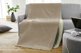 Beige Jacquard-Decke 150x200cm auf einem grauen Sofa drapiert, mit der Aufschrift 'Gemütlichmacher', in einem modernen Wohnzimmer mit Holzboden und Pflanzen im Hintergrund.