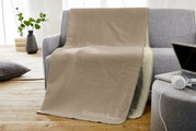 Beige Jacquard-Decke 150x200cm auf einem grauen Sofa drapiert, mit der Aufschrift 'Gemütlichmacher', in einem modernen Wohnzimmer mit Holzboden und Pflanzen im Hintergrund.