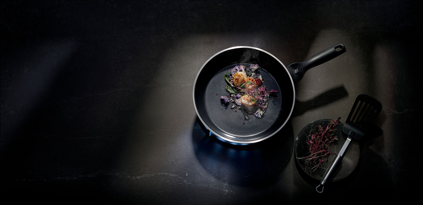 Pfanne CERADUR PROFI mit gebratenen Jakobsmuscheln und Gemüse, von oben fotografiert, neben einem Teller mit Kräutern und einem Pfannenwender.