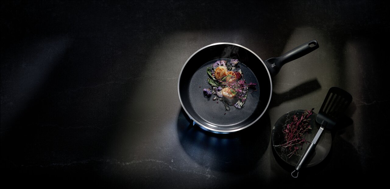 Pfanne CERADUR PROFI mit gebratenen Jakobsmuscheln und Gemüse, von oben fotografiert, neben einem Teller mit Kräutern und einem Pfannenwender.