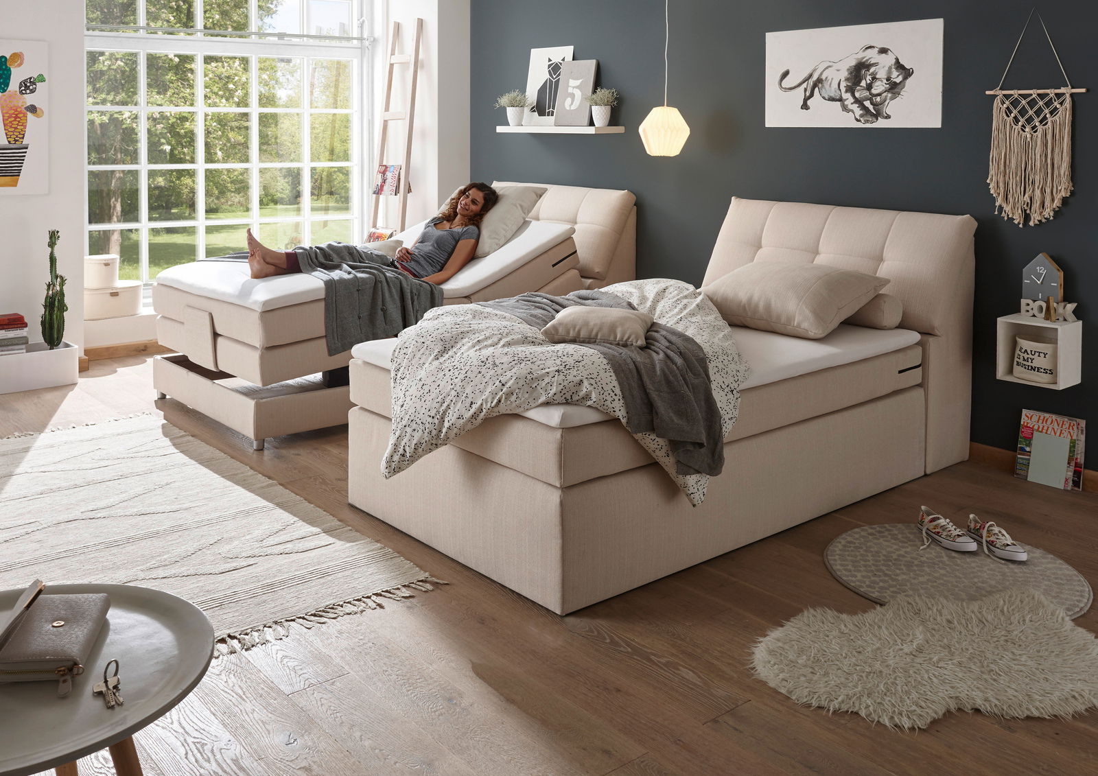 Zwei beige Boxspringbetten in einem modernen Schlafzimmer mit großen Fenstern, eines mit verstellbarem Kopfteil, seitliche Perspektive.