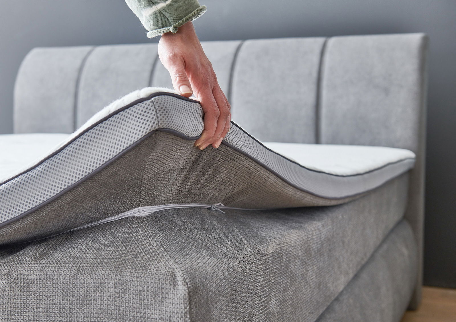 Nahaufnahme eines grauen Boxspringbetts mit einer Hand, die die Matratze anhebt, um die Struktur und den Bezug zu zeigen.