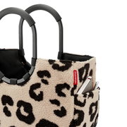 Nahaufnahme in Frontansicht einer beige-schwarzen Reisenthel Tragetasche aus Teddyfleece mit Leopardenmuster, schwarzen Griffen und Außenfach mit eingestecktem Smartphone und Stift.