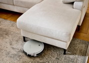 Beiger Stoff-Fußhocker einer Wohnlandschaft mit schwarzen Füßen, auf einem gemusterten Teppich; ein runder Saugroboter fährt darunter – Nahaufnahme aus leicht schräger Seitenansicht von vorne.