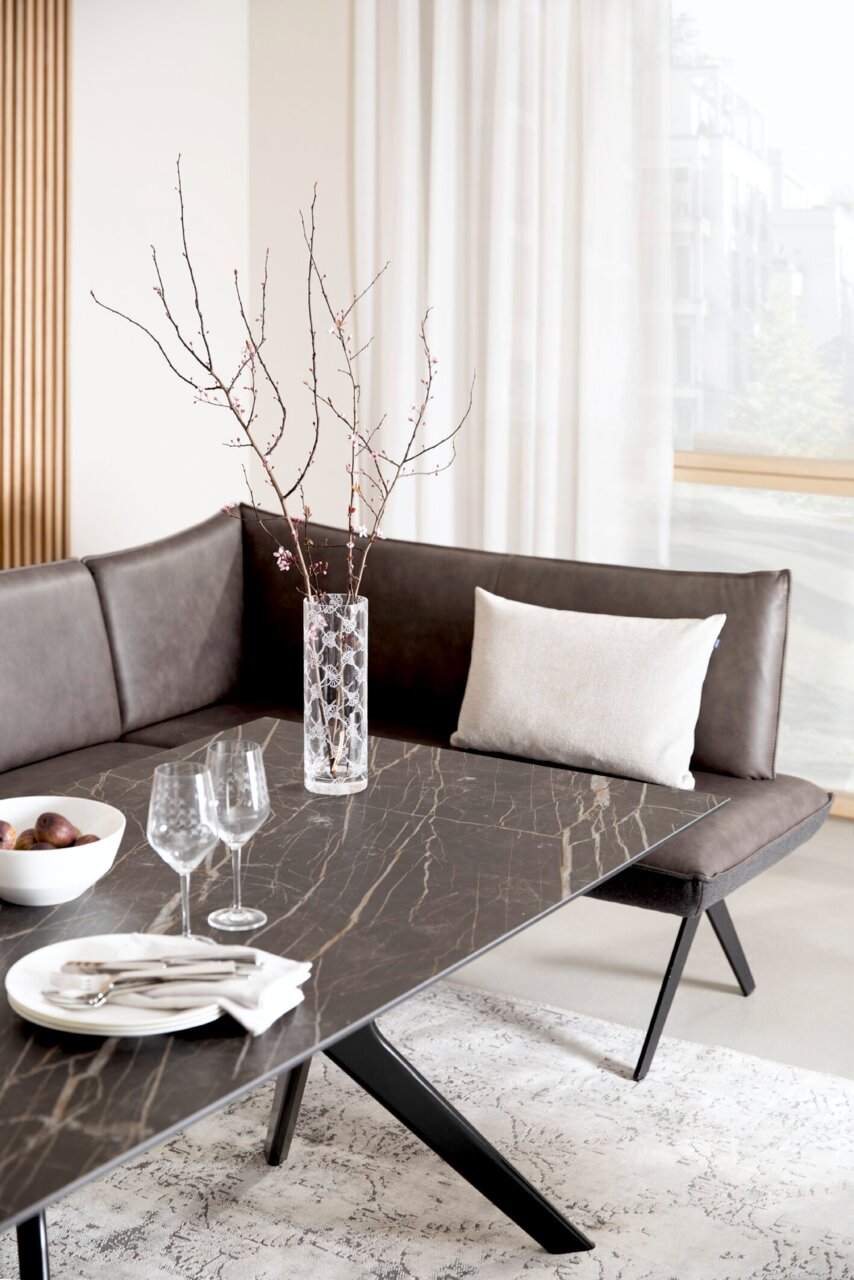 Moderne Eckbank in dunklem Leder mit dekorativem Kissen, fotografiert aus einer seitlichen Perspektive. Der Tisch hat eine elegante, dunkle Marmoroptik und ist mit Gläsern und einer Schale dekoriert.