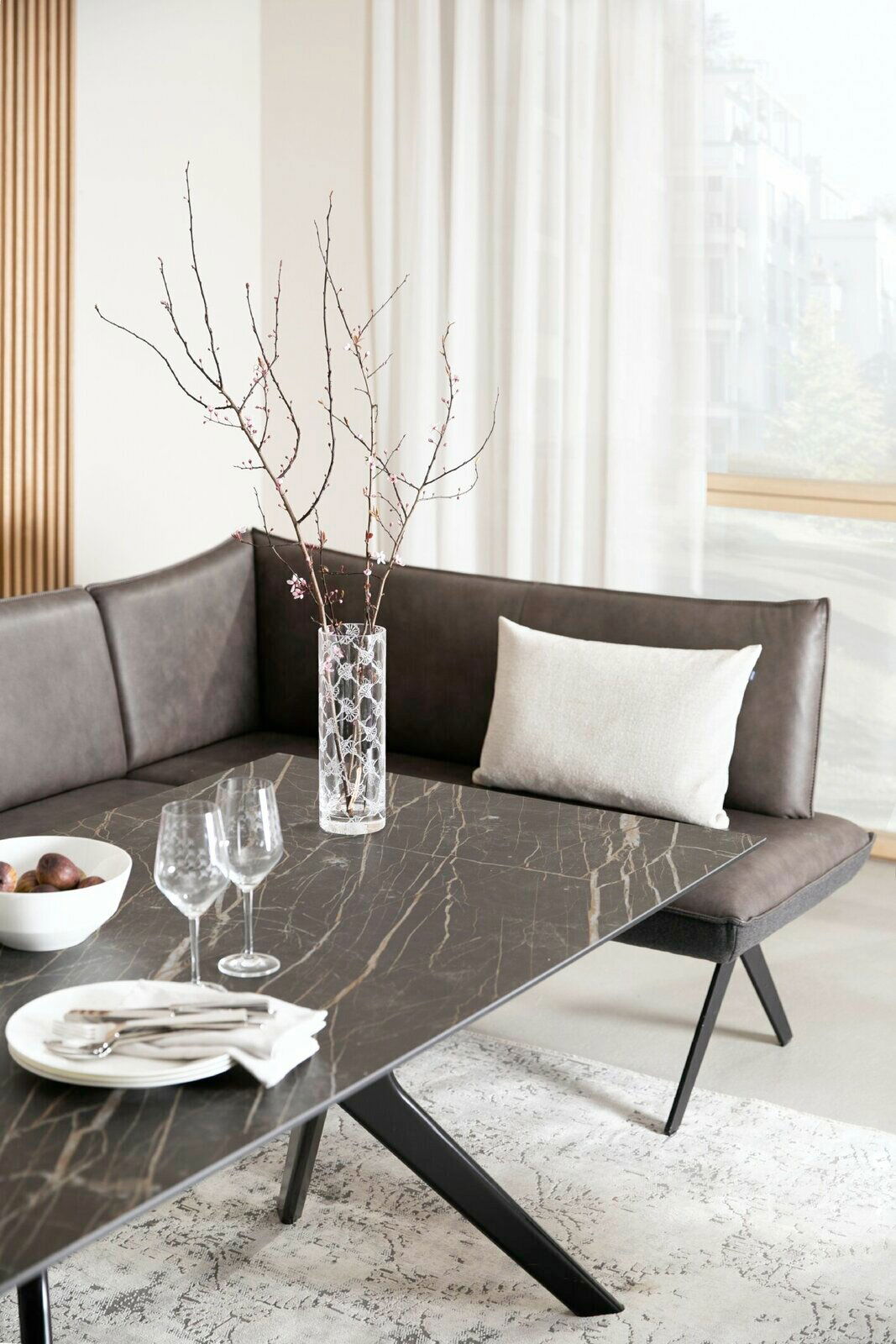 Moderne Eckbank in dunklem Leder mit dekorativem Kissen, fotografiert aus einer seitlichen Perspektive. Der Tisch hat eine elegante, dunkle Marmoroptik und ist mit Gläsern und einer Schale dekoriert.