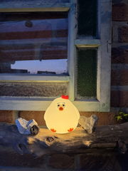 Niedliches Akku-Nachtlicht in Form eines Huhns, leuchtend auf einem Holzbrett vor einem Fenster, frontal betrachtet.