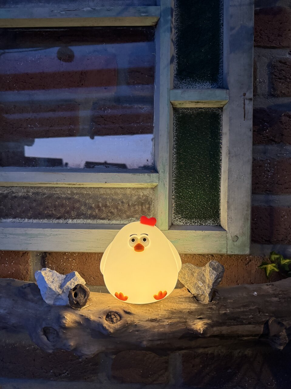 Niedliches Akku-Nachtlicht in Form eines Huhns, leuchtend auf einem Holzbrett vor einem Fenster, frontal betrachtet.