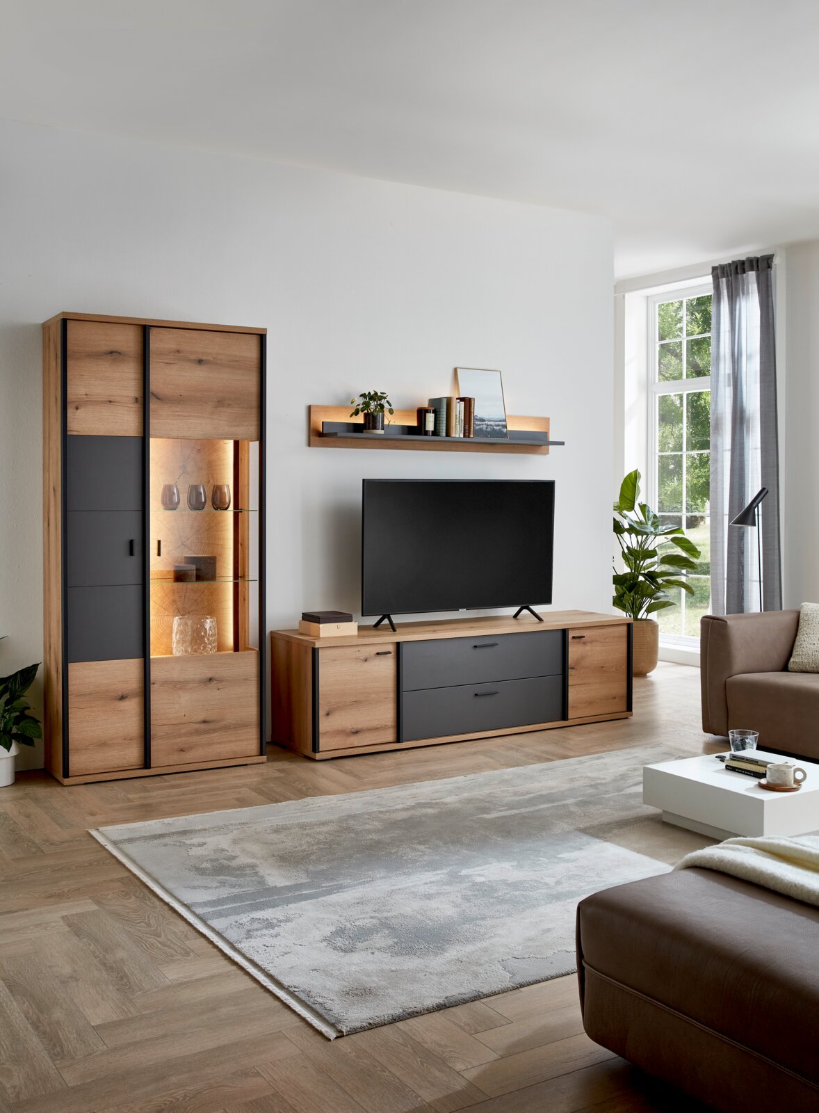 Modernes Wohnzimmer-Setup mit Wohnwand aus Holzdekor und anthrazitfarbenen Fronten, bestehend aus Vitrine mit Beleuchtung, TV-Lowboard und Wandboard, frontal in leichter Schrägansicht von rechts aufgenommen.