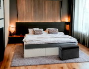Modernes Doppelbett mit dunkel gepolstertem Kopfteil, weißer Bettwäsche und grauem Hocker am Fußende in einem Schlafzimmer mit Nachttischen und warmen Lampenlicht, Frontansicht leicht erhöht.