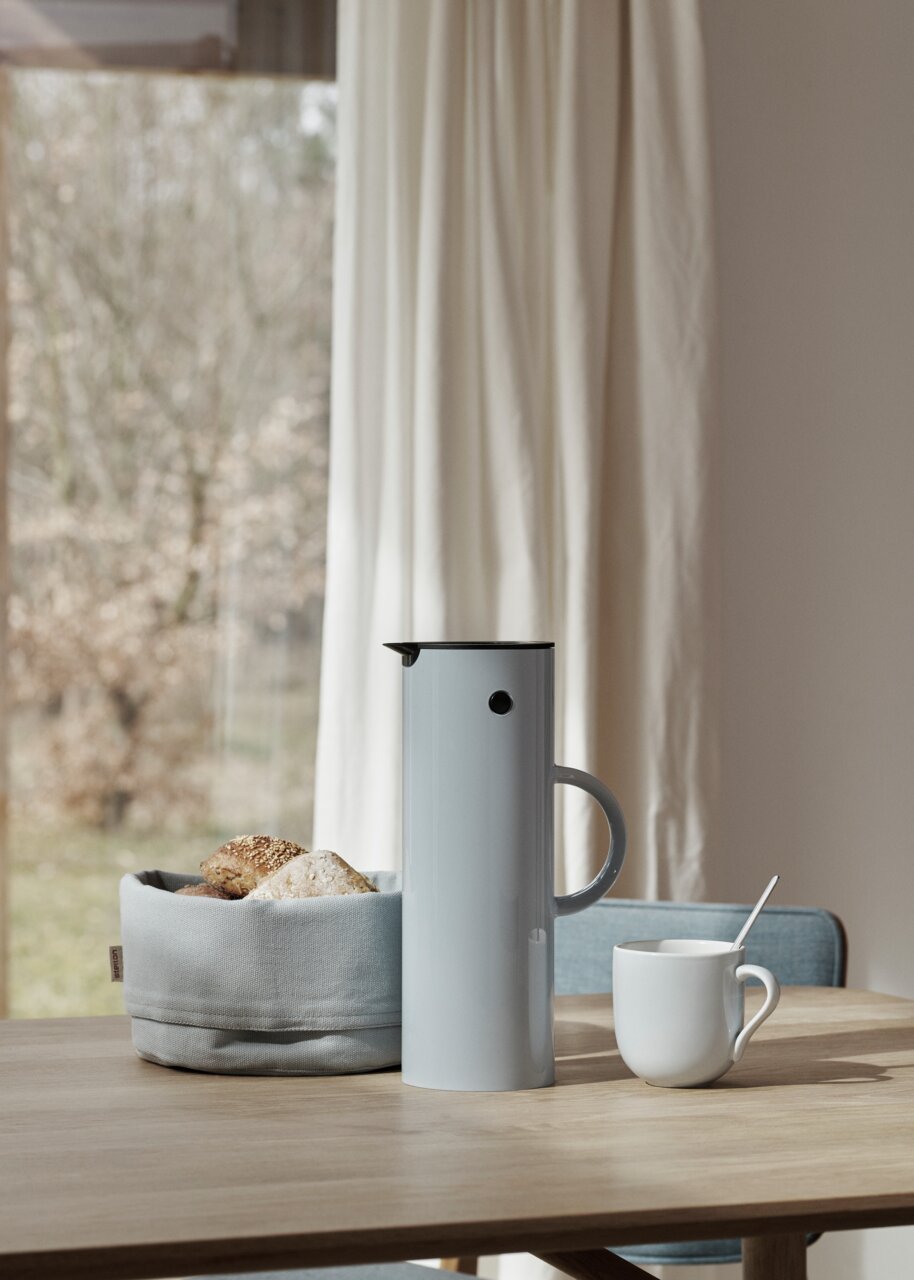 Stelton Isolierkanne 1l EM77 Elegante Isolierkanne in Wolkenfarbe auf einem Tisch, umgeben von einem Brotkorb und einer Tasse, seitliche Perspektive.