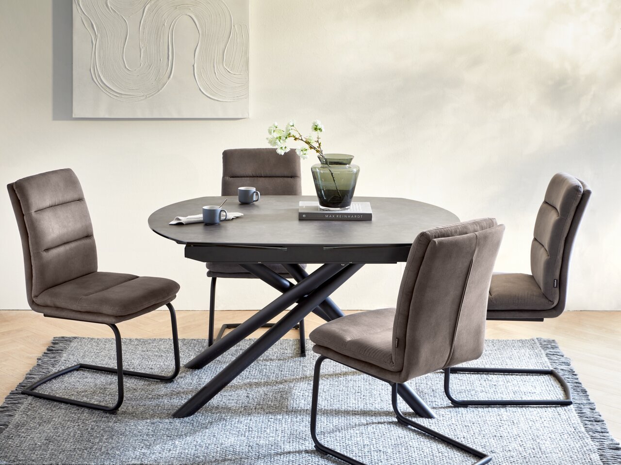 Ronde eettafel met zwart blad en gekruiste poten, omringd door vier gestoffeerde stoelen in een moderne eetkamer, gezien vanuit de zijkant.