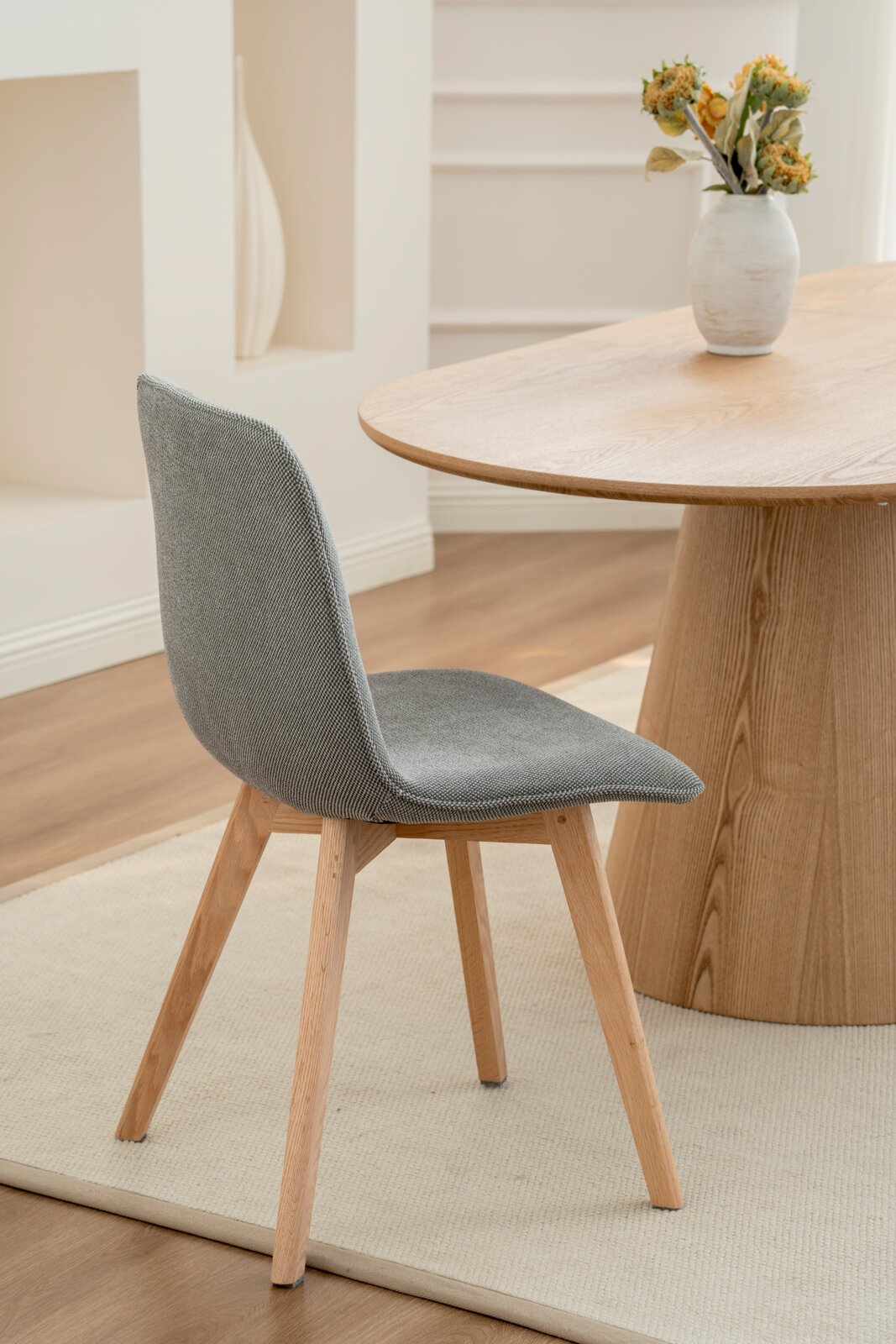 Moderner grauer Stuhl mit Holzbeinen, seitliche Perspektive, neben einem Holztisch in einem hellen Raum.
