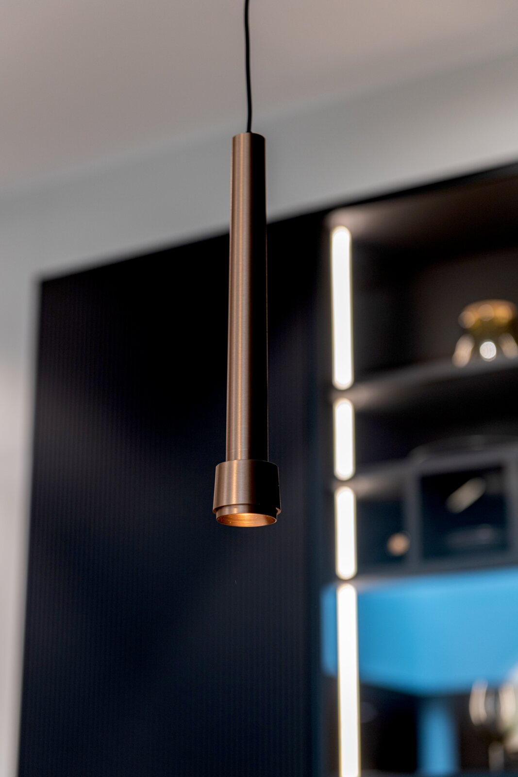 Schlanke, zylindrische Pendelleuchte aus gebürstetem Metall in Bronze/Kupferoptik mit schwarzem Kabel, Nahaufnahme aus leicht seitlicher Perspektive vor unscharfem modernem Innenraum