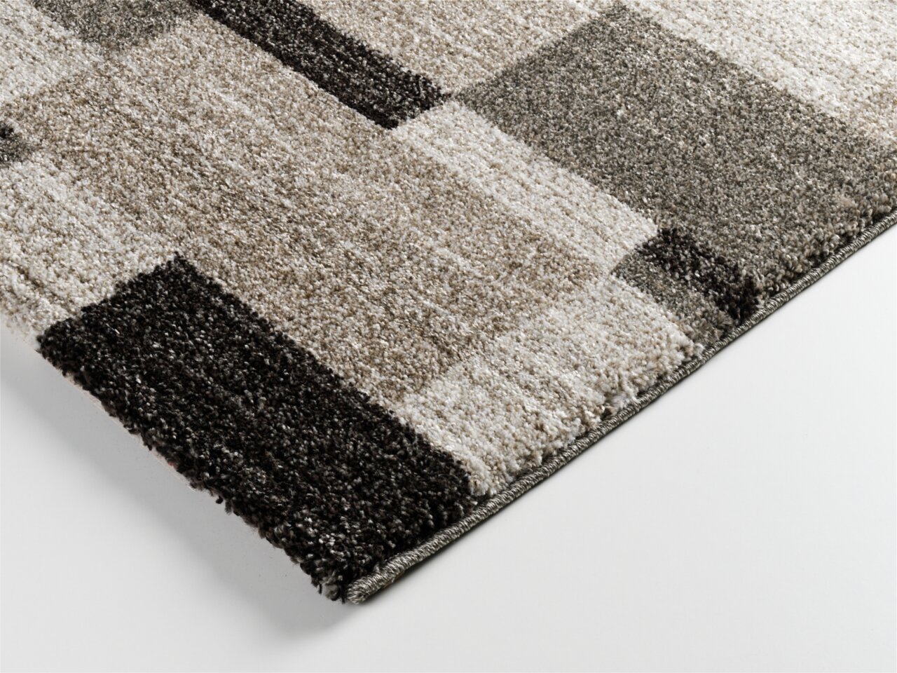 Nahaufnahme eines Hochflorteppichs Castle Beppo in rechteckigem Design mit verschiedenen Grautönen und geometrischem Muster, perspektivisch von der Seite aufgenommen.