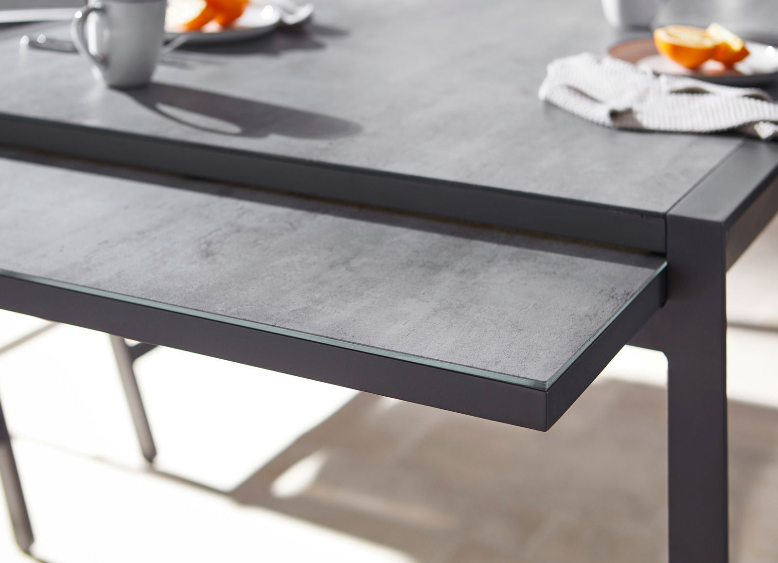 Nahaufnahme eines modernen Gartentisches mit grauer Steinoptik-Oberfläche und schwarzem Metallrahmen, seitliche Perspektive