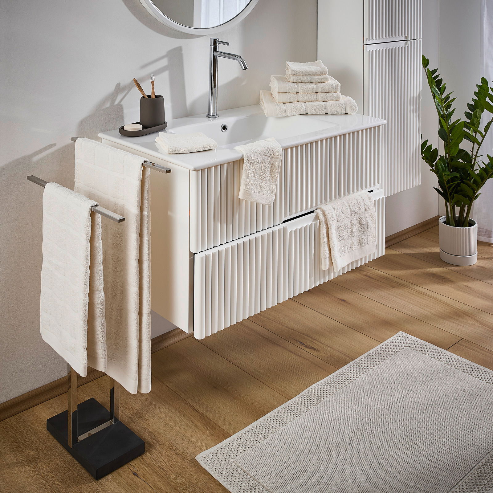 Badezimmer mit einem eleganten Frottierset in Cremefarbe, bestehend aus Handtüchern und Waschlappen, die auf einem modernen Waschbecken und Handtuchhalter arrangiert sind. Perspektive von der Seite.