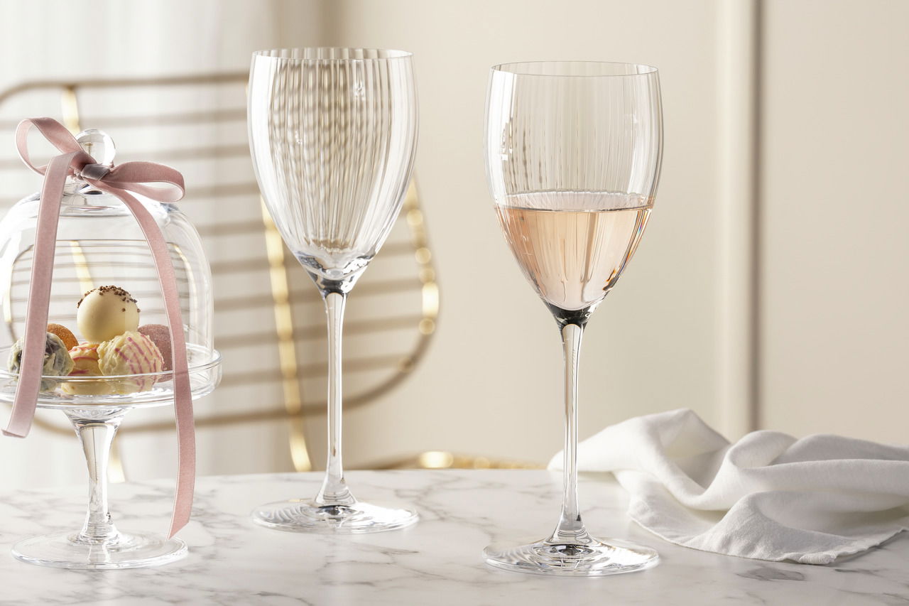 Leonardo Rieslingglas POESIA Zwei elegante Rieslinggläser der Serie POESIA mit feinen Rillen, eines gefüllt mit einem roséfarbenen Getränk, auf einem Marmortisch mit einer Glasglocke voller Pralinen und einem weißen Tuch im Hintergrund. Seitliche Perspektive.