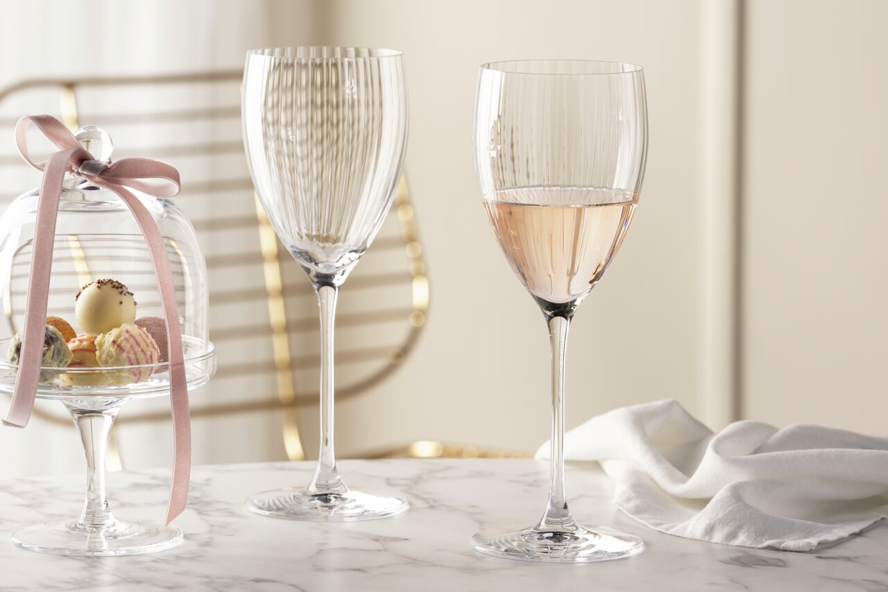 Zwei elegante Rieslinggläser der Serie POESIA mit feinen Rillen, eines gefüllt mit einem roséfarbenen Getränk, auf einem Marmortisch mit einer Glasglocke voller Pralinen und einem weißen Tuch im Hintergrund. Seitliche Perspektive.