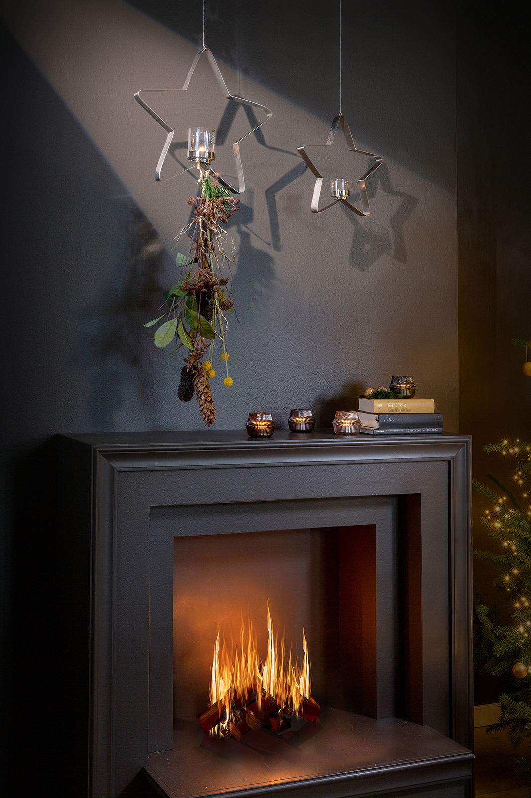 Zwei sternförmige Teelichthalter aus Metall hängen von der Decke, einer mit dekorativem Grün und Zapfen geschmückt, vor einem dunklen Hintergrund über einem modernen Kamin mit brennendem Feuer. Perspektive: Frontalansicht.
