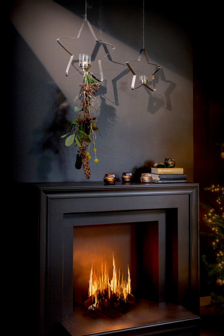 Zwei sternförmige Teelichthalter aus Metall hängen von der Decke, einer mit dekorativem Grün und Zapfen geschmückt, vor einem dunklen Hintergrund über einem modernen Kamin mit brennendem Feuer. Perspektive: Frontalansicht.