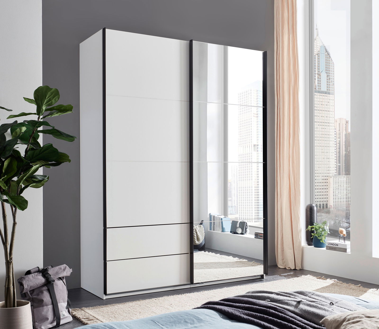 Moderner, zweiflügeliger Schwebetürenschrank mit Spiegel, in einem hellen Raum mit großen Fenstern, aus der Perspektive von schräg vorne fotografiert.