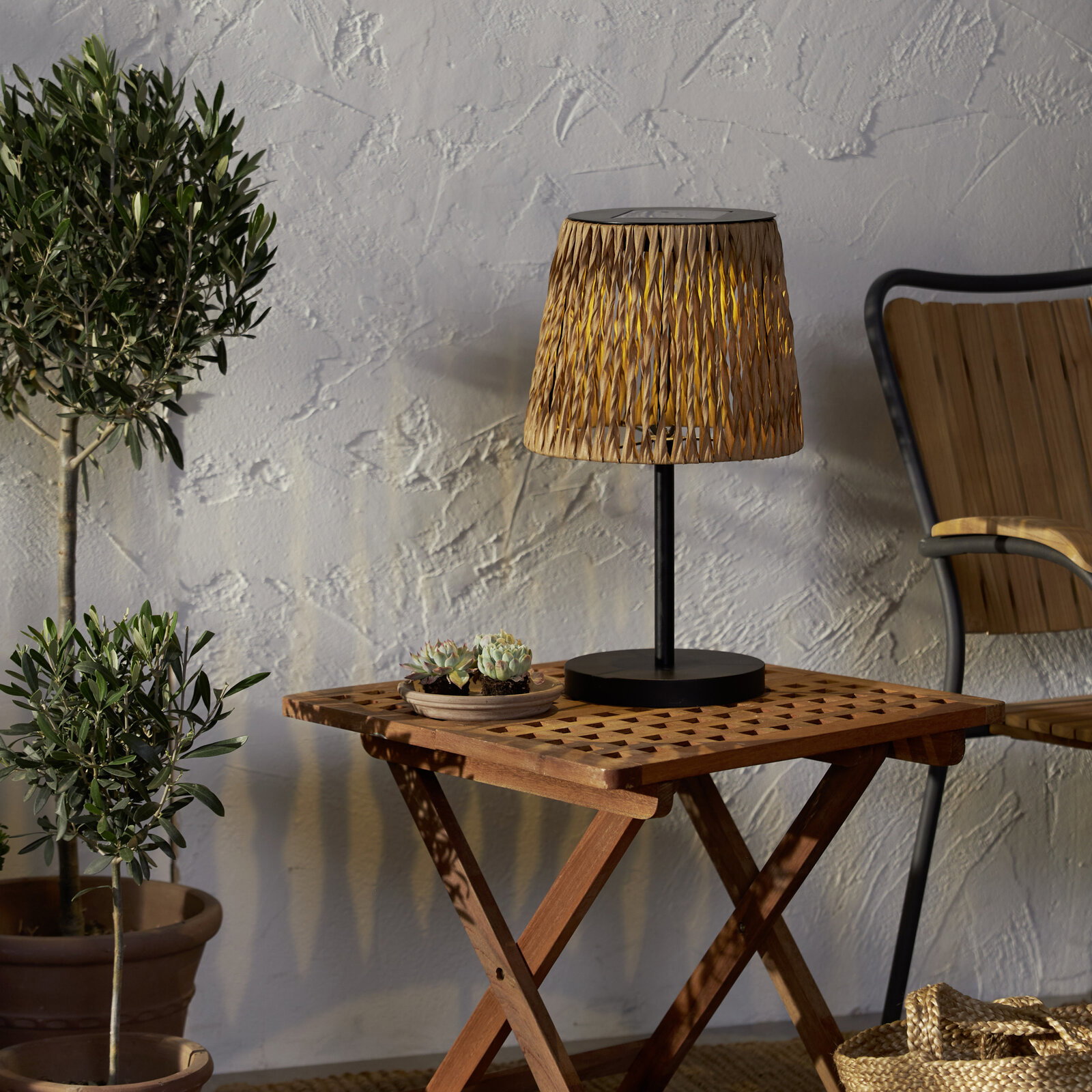 Dekorative Tischlampe mit geflochtenem Schirm in Naturton und schwarzem Fuß auf einem klappbaren Holztisch, seitliche Perspektive im Outdoor-Setting mit Pflanzen und Stuhl im Hintergrund