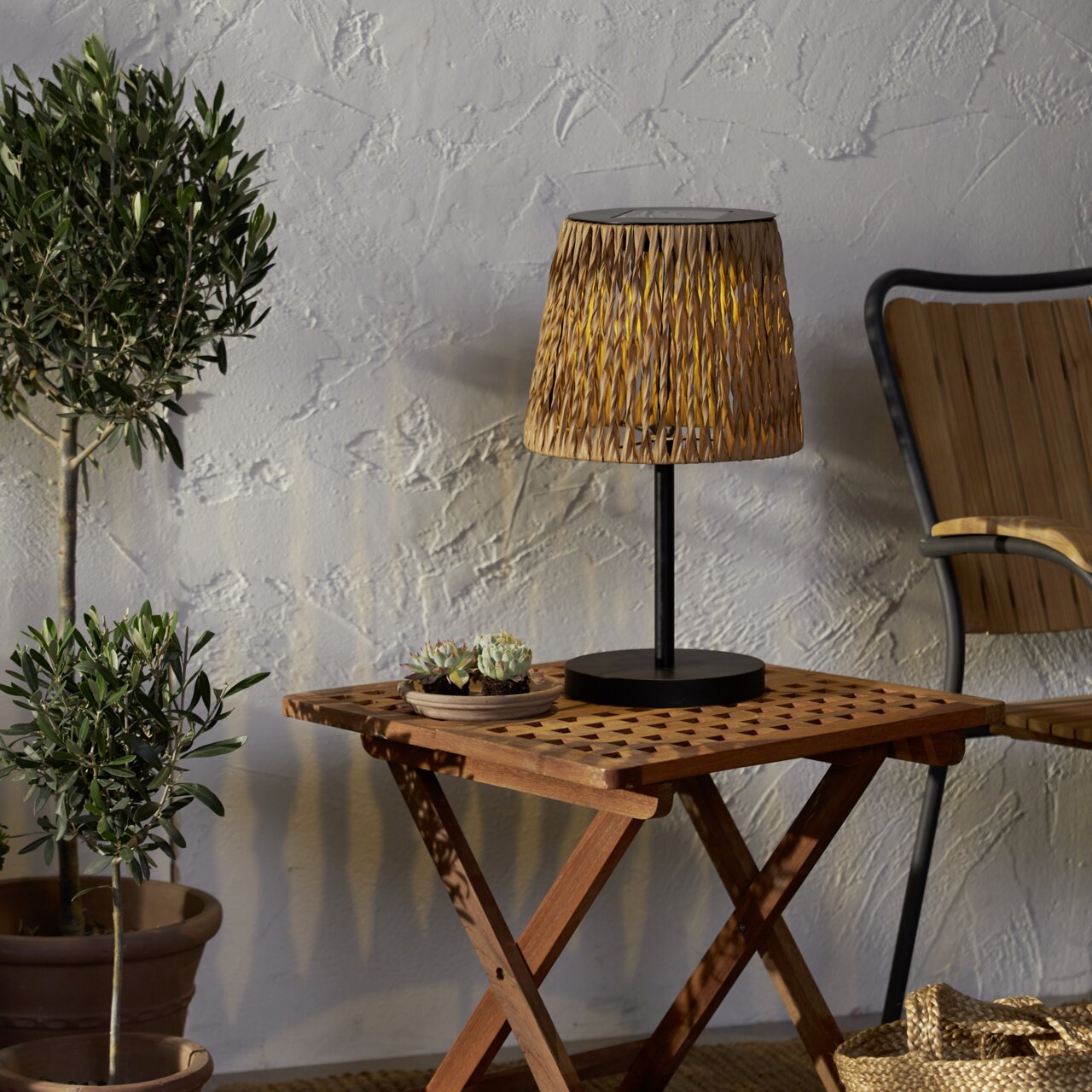 Dekorative Tischlampe mit geflochtenem Schirm in Naturton und schwarzem Fuß auf einem klappbaren Holztisch, seitliche Perspektive im Outdoor-Setting mit Pflanzen und Stuhl im Hintergrund