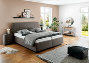 Modernes Polsterbett in einem stilvoll eingerichteten Schlafzimmer, seitliche Perspektive mit Nachttisch, Pflanzen und Dekoration.