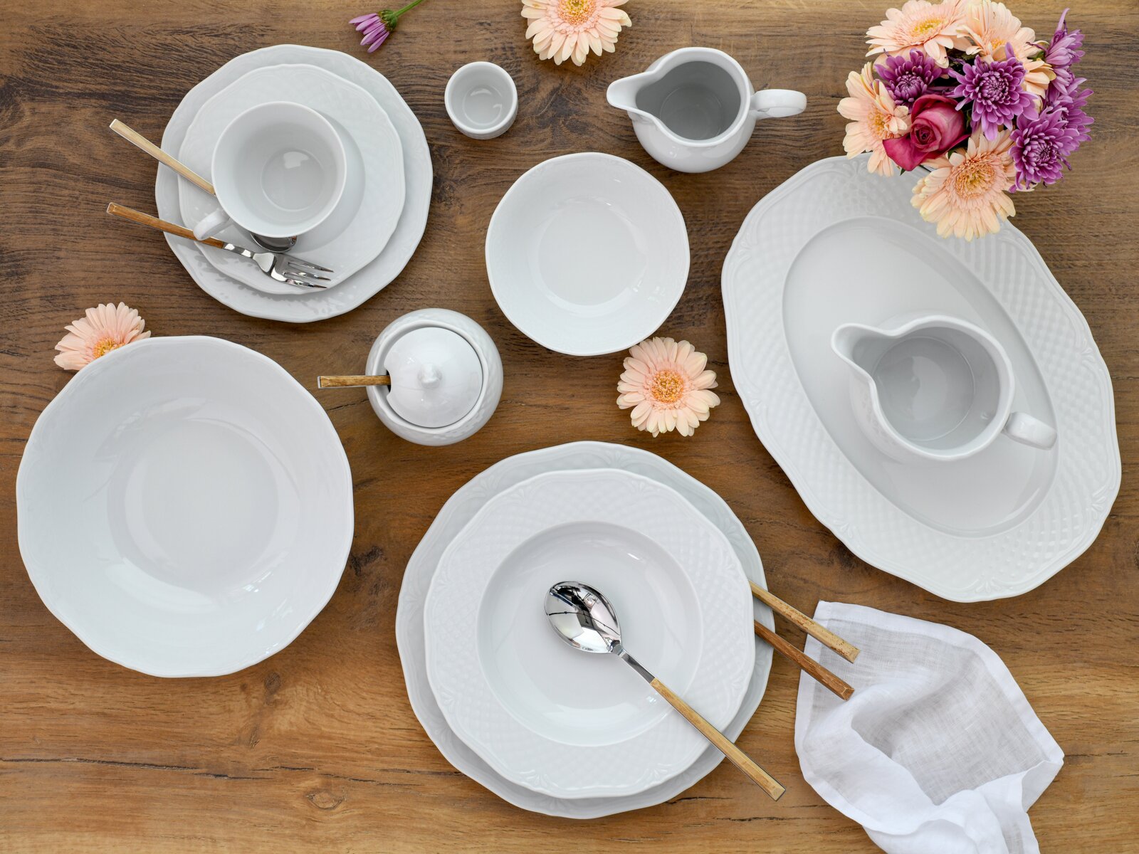 Draufsicht auf ein elegantes weißes Porzellan-Geschirrset mit Tellern, Schalen, Tasse, Milchkännchen und Zuckerdose auf einem Holztisch, dekoriert mit Blumen und Besteck.