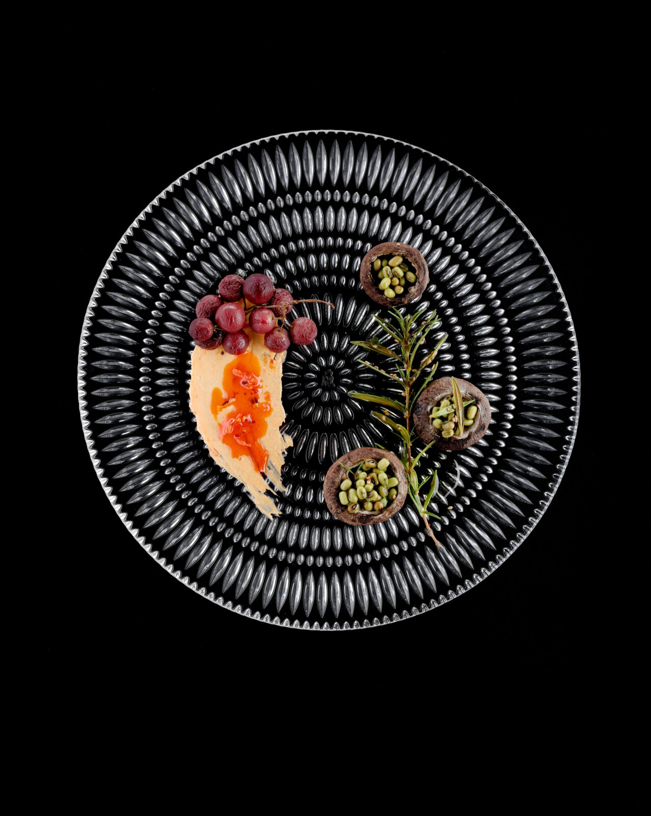 Ronde zwarte designbord met een reliëfachtig patroon, gefotografeerd van bovenaf, met daarop een romige spread met rode saus, druiven, een takje rozemarijn en kleine schaaltjes met groene peulvruchten
