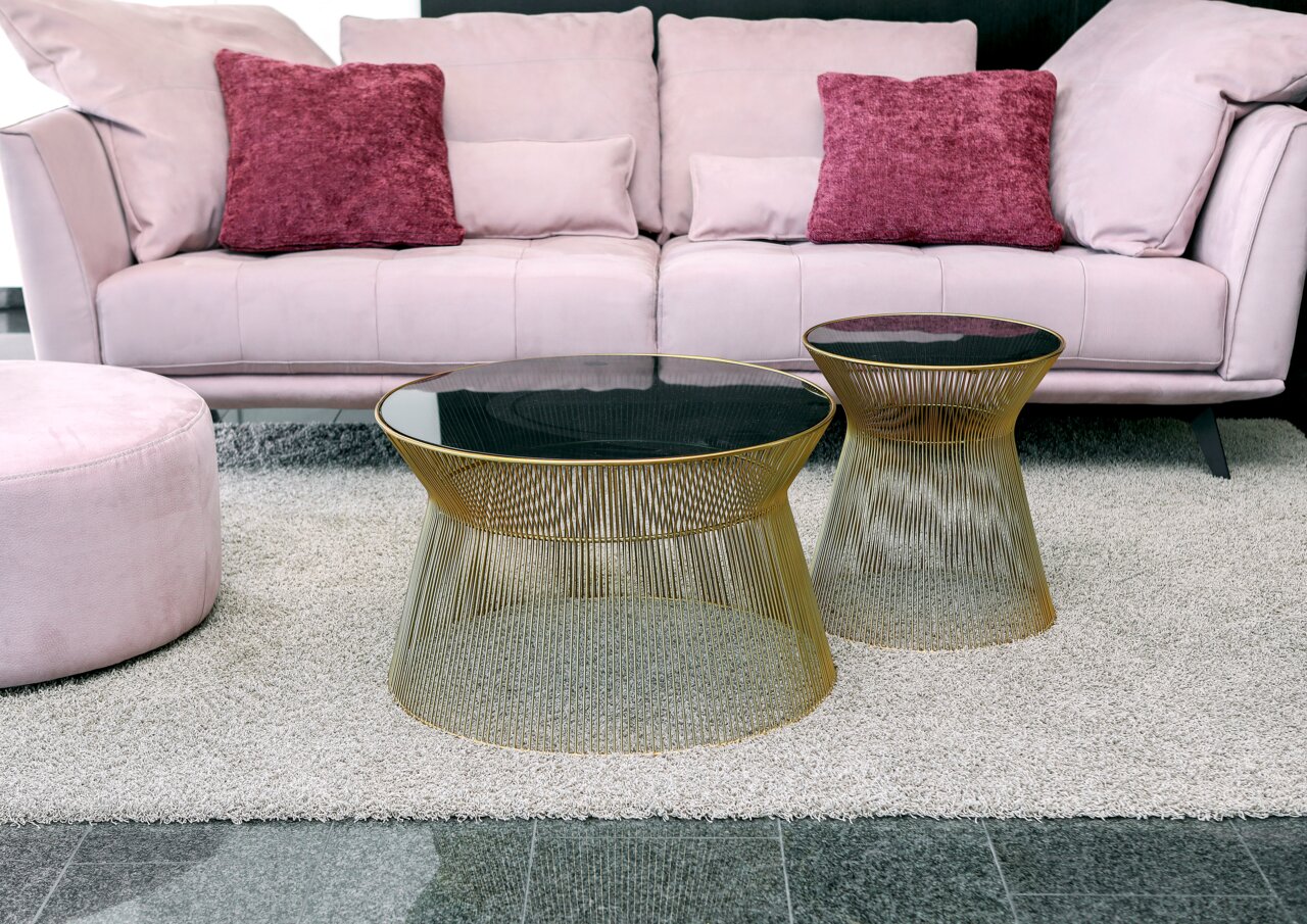 Moderner Couchtisch und Beistelltisch mit goldenen Drahtgestellen und schwarzer Glasplatte, fotografiert aus einer leicht erhöhten Perspektive vor einem rosa Sofa.