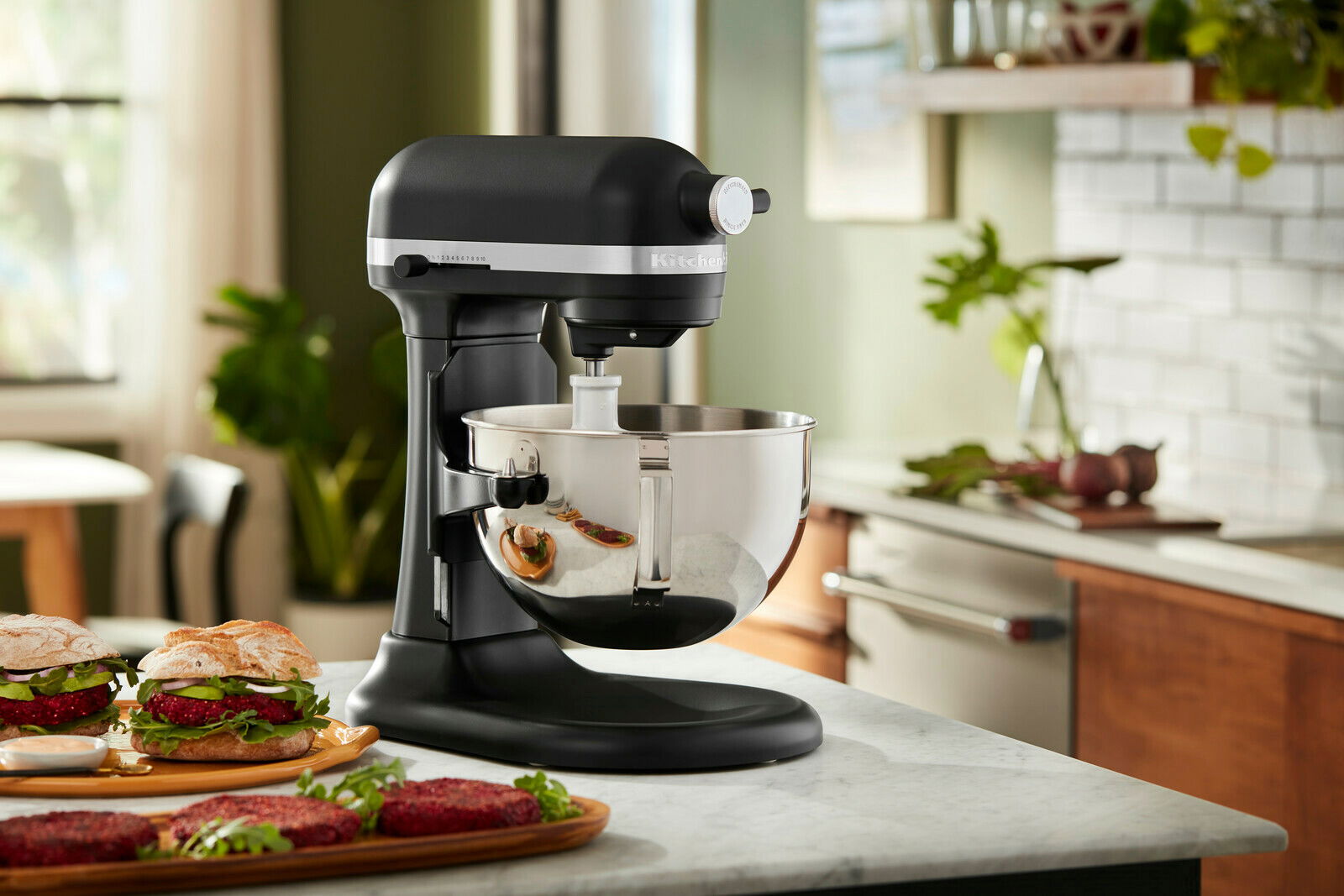 Schwarze Küchenmaschine Heavy Duty 5,2L auf einer Küchenarbeitsplatte, seitliche Perspektive, mit einer Edelstahlschüssel und Burgern im Vordergrund.