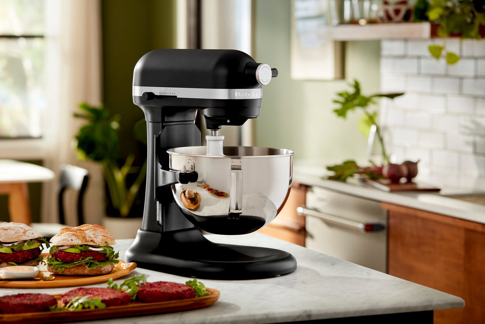 Schwarze Küchenmaschine Heavy Duty 5,2L auf einer Küchenarbeitsplatte, seitliche Perspektive, mit einer Edelstahlschüssel und Burgern im Vordergrund.