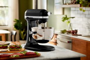 Schwarze Küchenmaschine Heavy Duty 5,2L auf einer Küchenarbeitsplatte, seitliche Perspektive, mit einer Edelstahlschüssel und Burgern im Vordergrund.