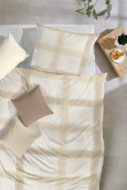 Draufsicht auf ein Bett mit hell gemusterter Bettwäsche in Beige/Creme und dekorativen Kissen; daneben ein Holznachttisch mit Pflanze und Gebäck.