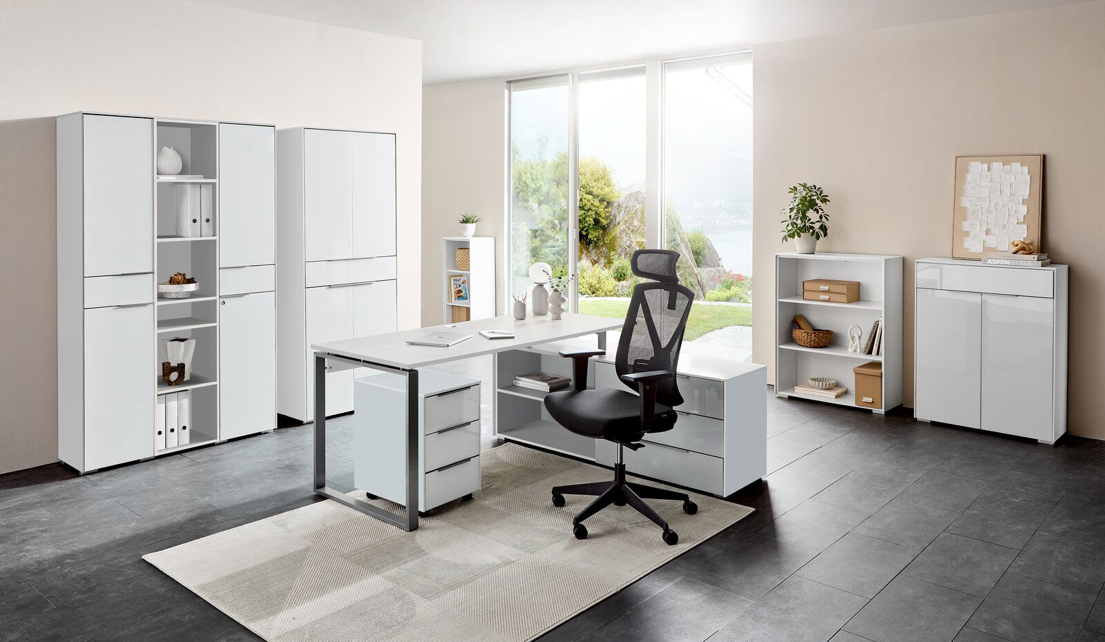 Moderne Büromöbelkombination mit Schreibtisch, Rollcontainer und Aktenschränken in einem hellen Raum mit Blick auf den Garten, seitliche Perspektive.