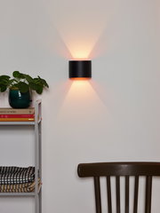 Moderne Wandleuchte XIO in schwarzem Zylinderdesign, an einer weißen Wand montiert, mit warmem Licht nach oben und unten. Perspektive von vorne.