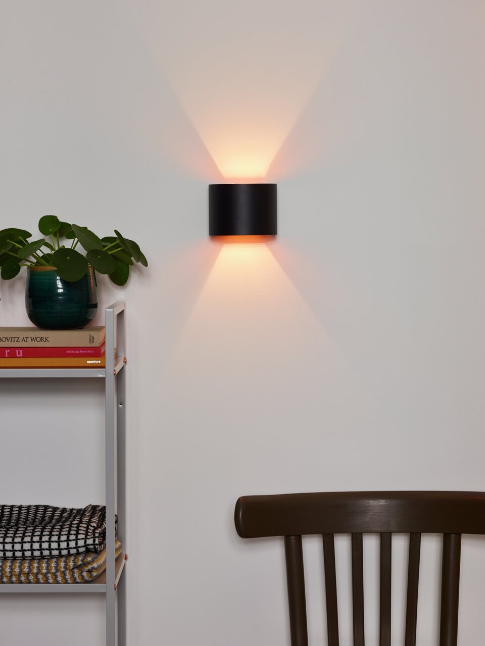 Moderne wandlamp XIO in zwart cilinderontwerp, gemonteerd op een witte muur, met warm licht naar boven en beneden. Aanzicht van voren.