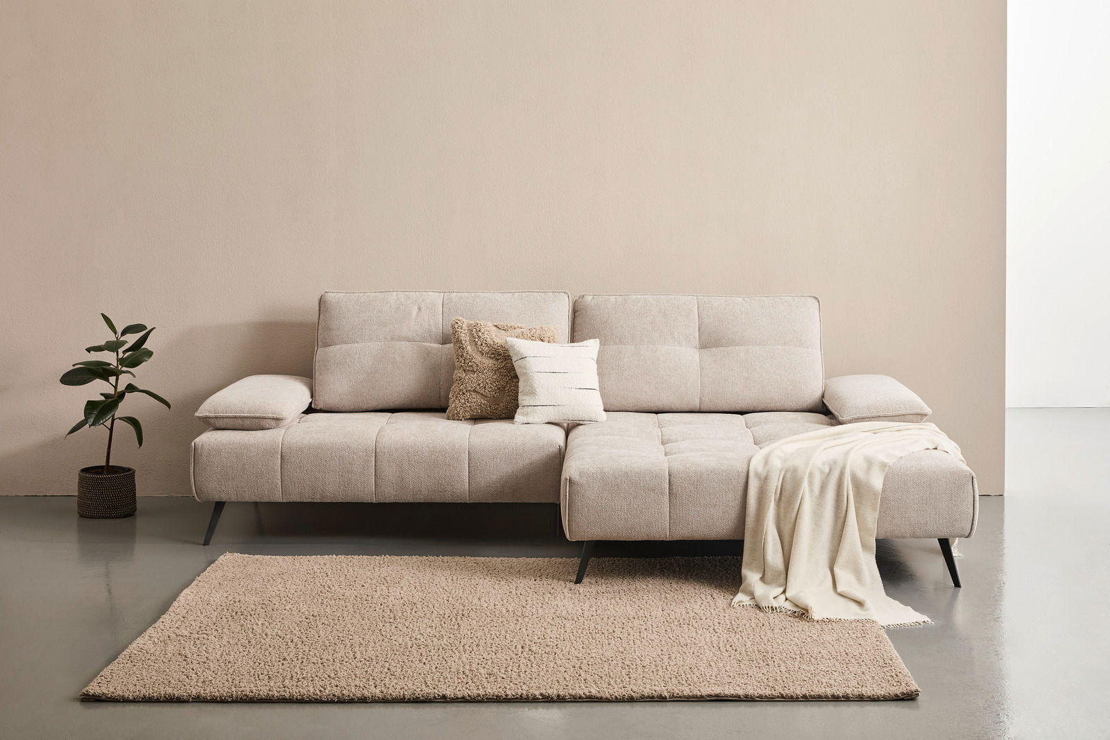Beige Longchairkombination mit zwei Kissen und einer Decke, frontal fotografiert, auf einem Teppich neben einer Pflanze.