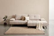 Beige Longchairkombination mit zwei Kissen und einer Decke, frontal fotografiert, auf einem Teppich neben einer Pflanze.