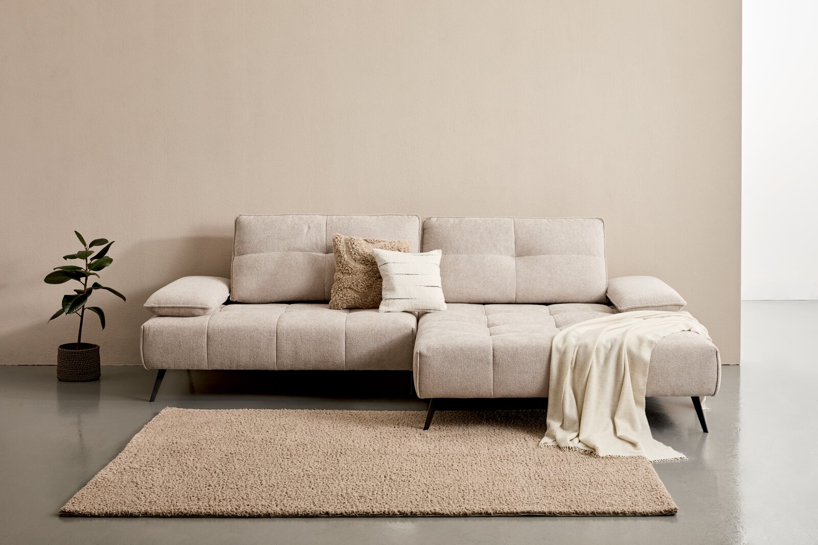 Beige Longchairkombination mit zwei Kissen und einer Decke, frontal fotografiert, auf einem Teppich neben einer Pflanze.