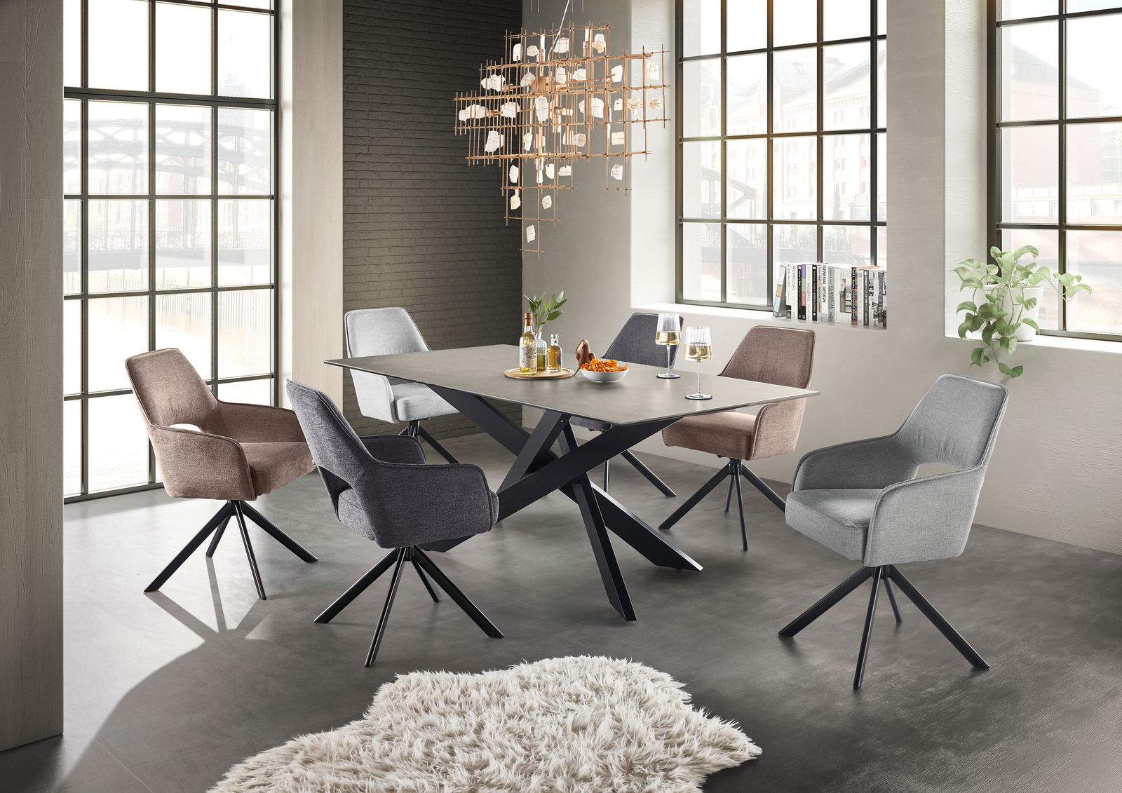 Moderner Esstisch mit X-förmigem Metallgestell und grauer Tischplatte, umgeben von sechs gepolsterten Stühlen in verschiedenen Farben, in einem hellen Raum mit großen Fenstern, aus seitlicher Perspektive.