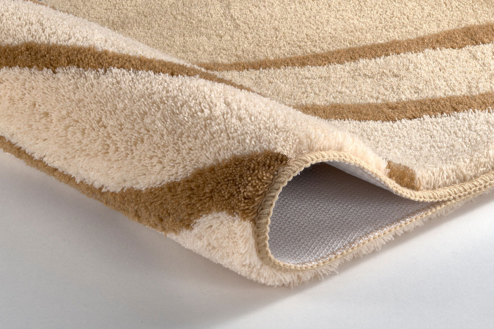 Nahaufnahme eines beigen und braunen Badteppichs mit weicher Textur, aufgerollte Ecke zeigt die rutschfeste Unterseite.