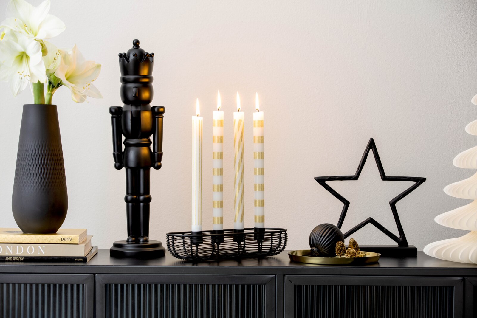 Frontalansicht eines stilvollen Weihnachtsarrangements in Schwarz, Weiß und Gold mit Nussknacker, vier brennenden Stabkerzen, Stern-Dekoration und Blumenvase auf einem schwarzen Sideboard.