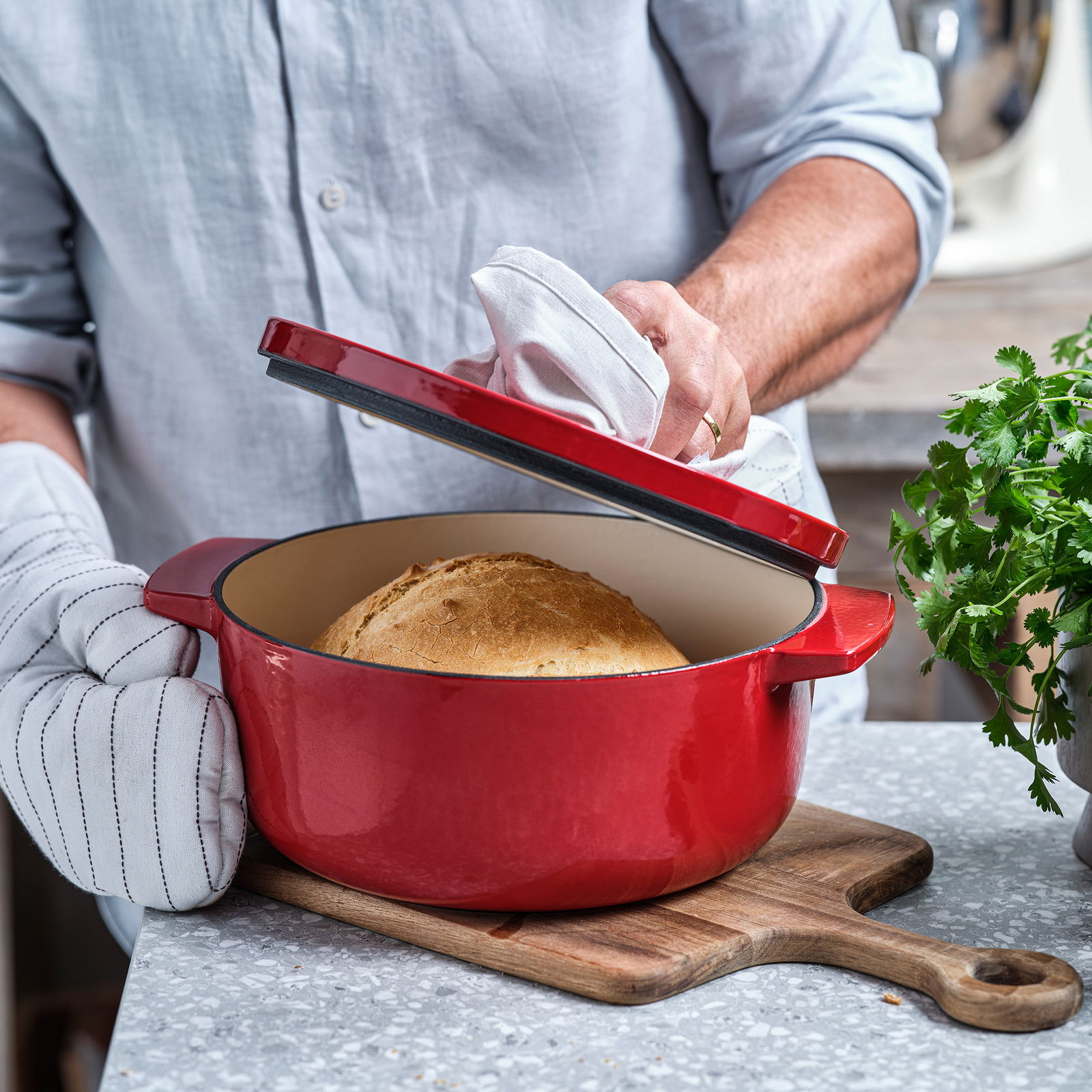 Roter runder Gusseisen-Bräter von KitchenAid mit Brot, geöffnet von einer Person mit Topfhandschuhen, seitliche Perspektive.